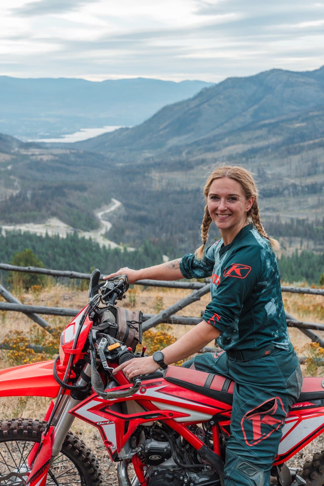 A smiling Dusthunniez rider with her bike at a mountain overlook, valley and lake in the background.
