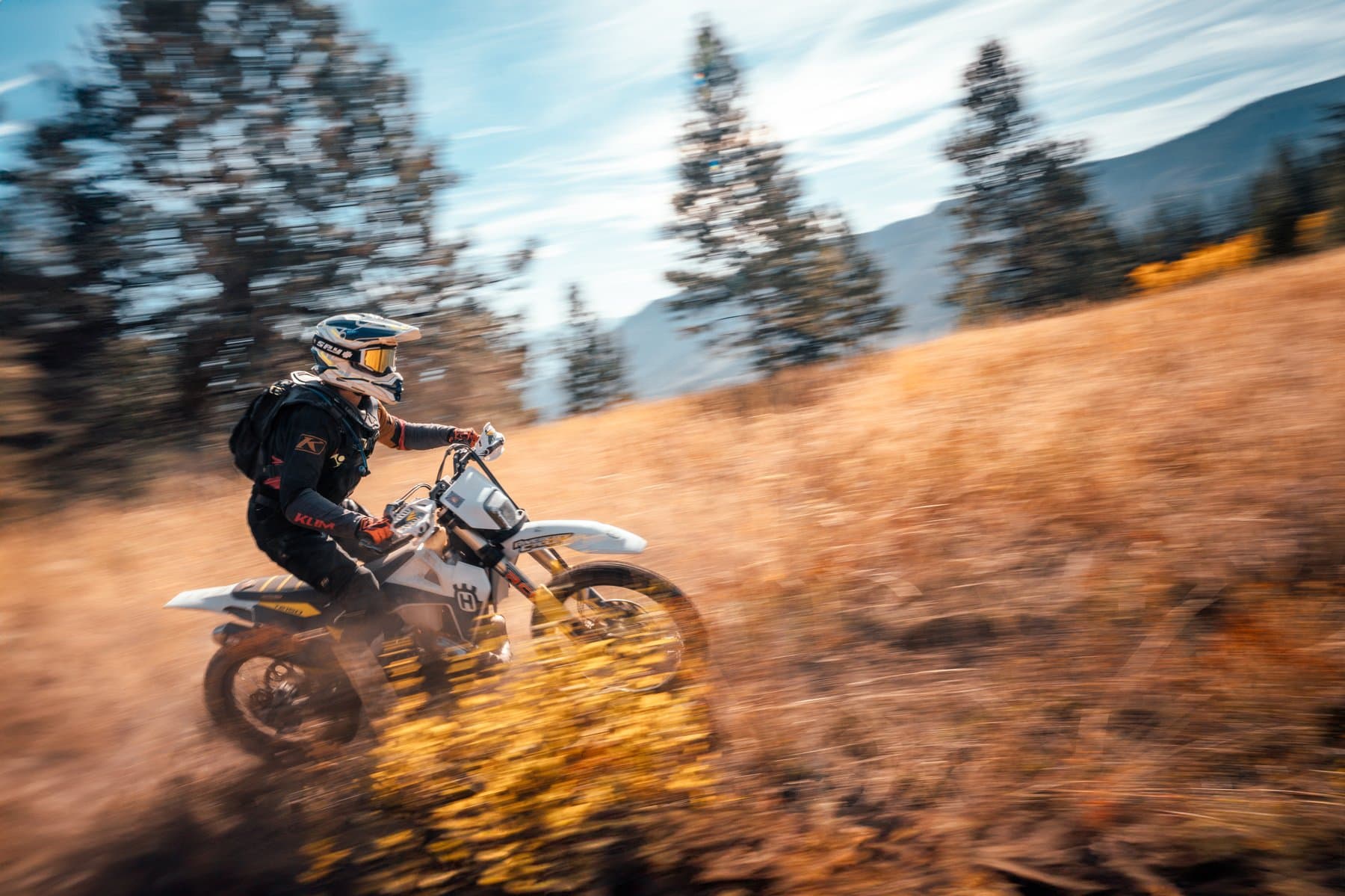 A Dusthunniez rider carves through a golden autumn field with mountains behind, motion-blurred panning shot.