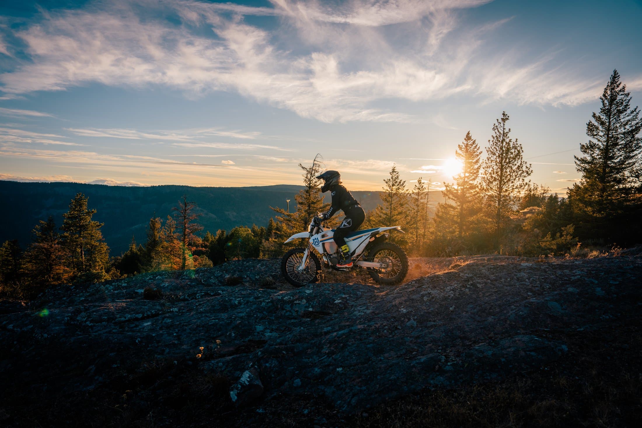 A lone rider silhouetted on a rocky ridge at sunset, sweeping BC forest and mountains behind her.